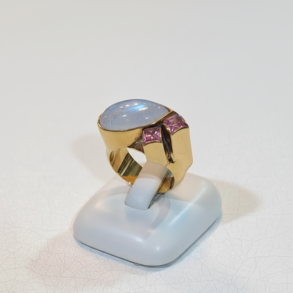 Gold ring with a large gemstone and pink stones on a white stand against a white background