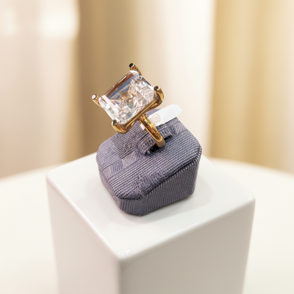 Gold ring with a large diamond on a textured gray stand against a neutral background