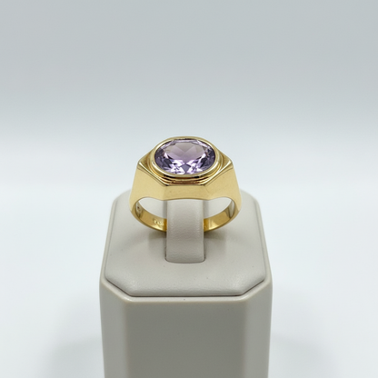 Gold ring with a purple gemstone on a white stand against a light gray background