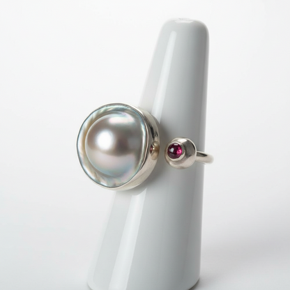 Silver ring with a large pearl and smaller red gemstone on a white stand.